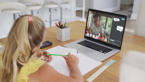 Schoolgirl-using-laptop-for-online-lesson-at-home,-with-female-teacher-and-webchat-on-screen