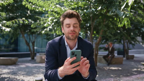 un hombre de negocios sonriente enviando mensajes de texto por teléfono móvil sentado en la plaza verde solo de cerca.
