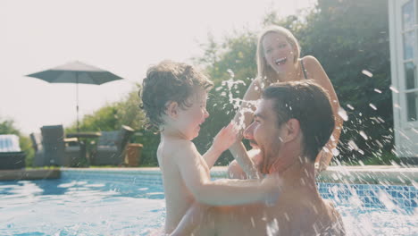 Familia-Con-Un-Hijo-Pequeño-Divirtiéndose-En-Las-Vacaciones-De-Verano-Jugando-Y-Chapoteando-En-La-Piscina-Al-Aire-Libre