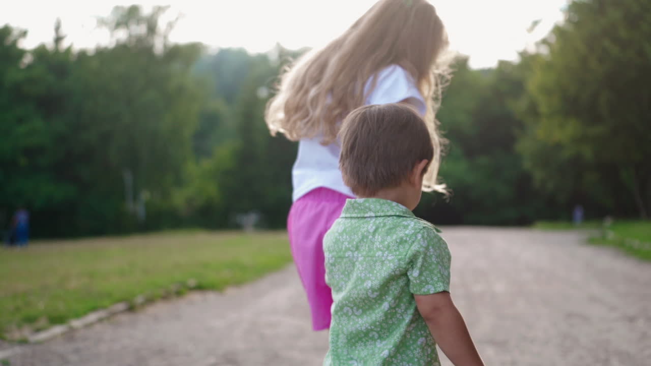 Sad boy going home with a little girl outdoors. Back view of two children holding hands and walking in the park in summer.