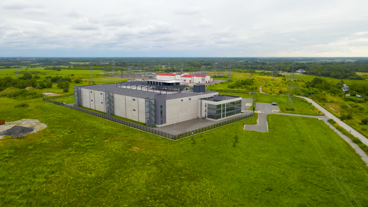 Aerial orbiting shot of a large and modern gray or grey colored data centre storage facility and a power station behind it with electrical lines during summer cloudy day in the countryside. roads too