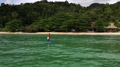 low angel aerial drone tracking shot of asian man exercising on a sup paddle board in turquoise tropical clear waters, with beach, small bungalow beach resort and coastline in thailand