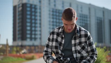 man in checkered shirt sits outdoors pulling out medication from shirt pocket while seated with soft blur of tall urban building and greenery in background during calm sunny day