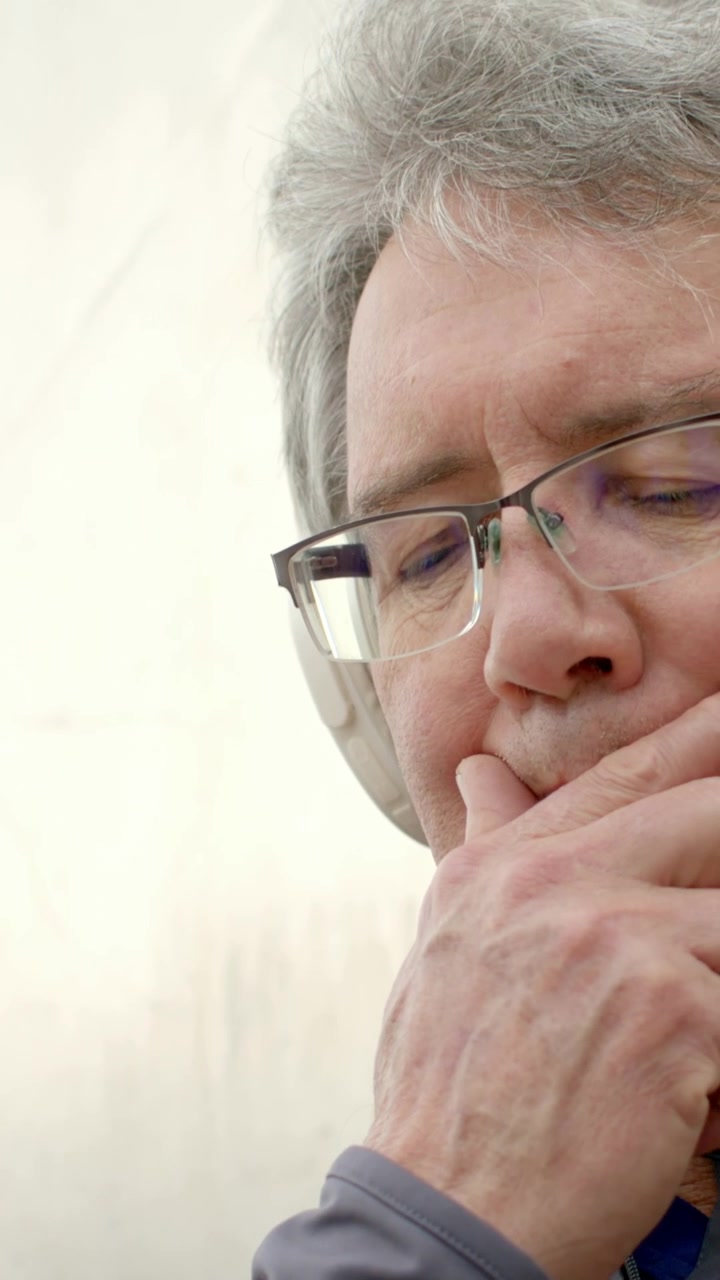 Vertical video: Looking downward, man glasses headphones bringing hand to lips in portrait thinking