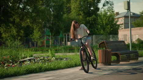 cyclist mounts bicycle near vibrant garden, preparing for ride surrounded by lush greenery, colorful flower beds, and sunlight, background includes wooden bench, trees, and serene park setting