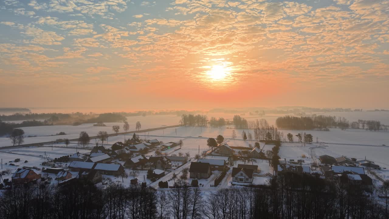 Praktfullt luftfoto av en snødekket landsby under en levende solnedgangshimmel. Nysnø dekker hustak og jorder, og fremkaller en rolig vinterstemning. Ideell for arkivbruk.