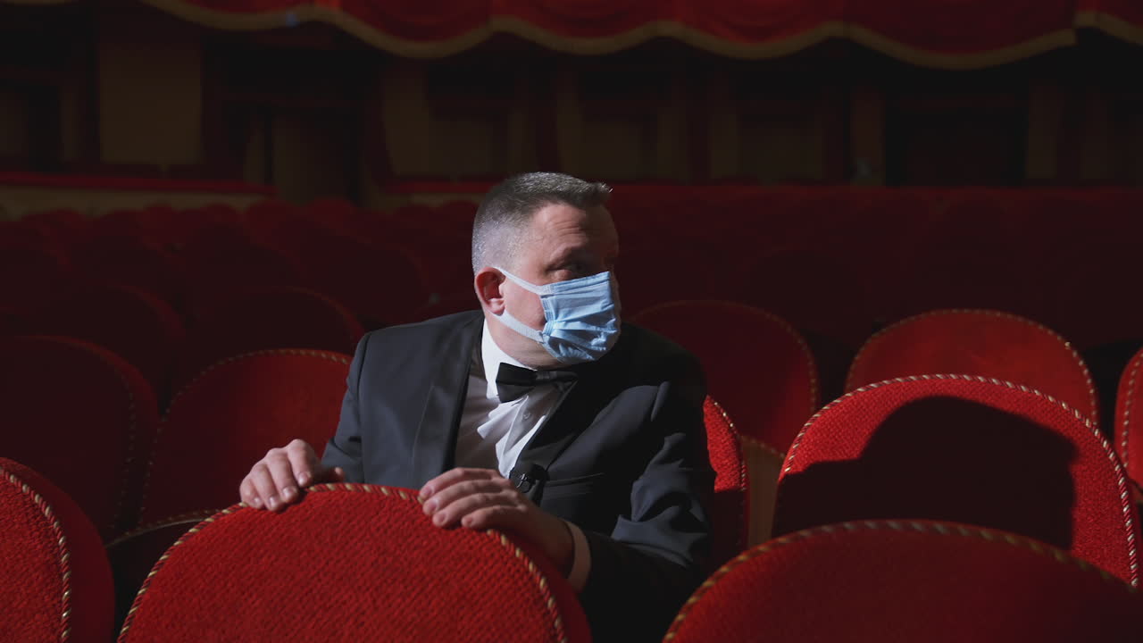 Theater after pandemic of covid-19. Man in medical mask sits alone in empty hall with red seats