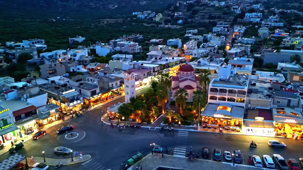 Animada plaza costera en Elounda con iglesia iluminada, tiendas y tráfico a la hora azul, vista desde un dron