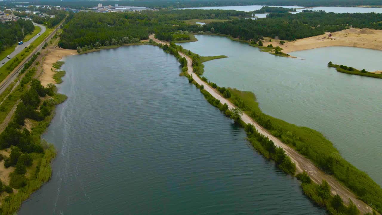 Aerial drone footage revealing a large sand quarry mine with two artificial lakes surrounding a minging gravel road leading to the sand dunes. Mining equipment and machines visible, highway on left