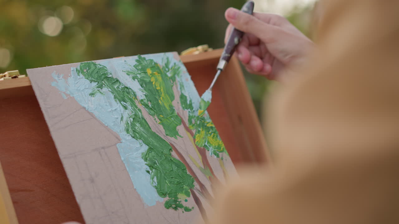 Utomhuskonstnär som målar träd-sketch på canvas, närbild av händer som applicerar grön och blå akryl med palettkniv i trädgårdsmiljö, höstljus, lugnt fokus, plein air-teknik