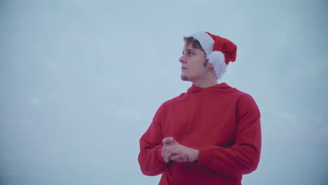 thoughtful man in red hooded jacket and santa hat looking away against sky