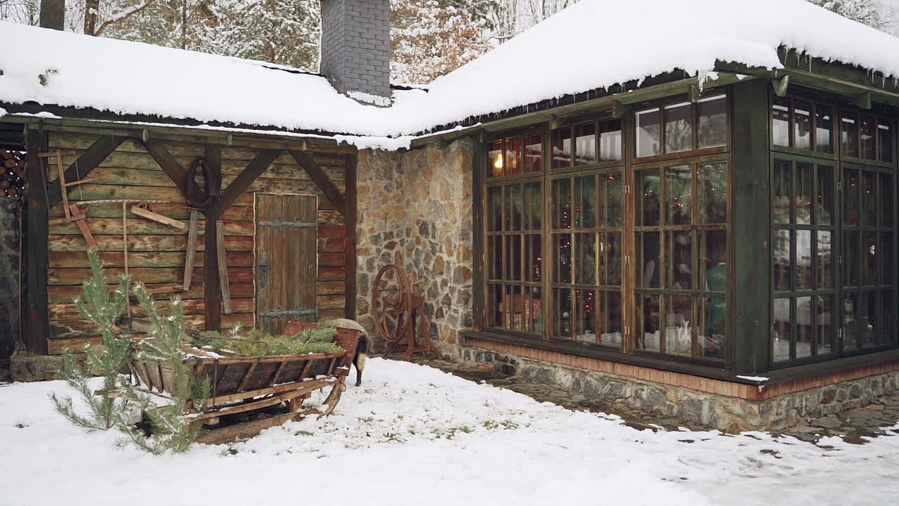 Beautiful cottage on the snowy background outdoors. Wooden sledge and a nice sheep standing in the street near the cozy building in the forest in winter.