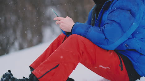 male snowboarder sitting on the snow taking photos on the phone beautiful scenery of the resort for social networks