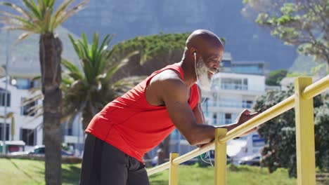 tired senior african american man wearing headphones using smartphone while leaning on railing of pr