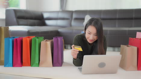 a young attractive woman lies on the floor surrounded by colorful shopping bags as she inputs her credit card number to her laptop