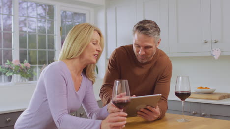 mature couple at home in kitchen using digital tablet to book show, vacation or make purchase and celebrating by drinking red wine together - shot in slow motion