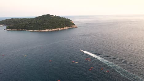 Aerial-view-of-the-Lokrum-Island-and-a-group-of-tourists-on-a-kayak-tour-and-a-boat-passing-by,-Dubrovnik-,-Croatia