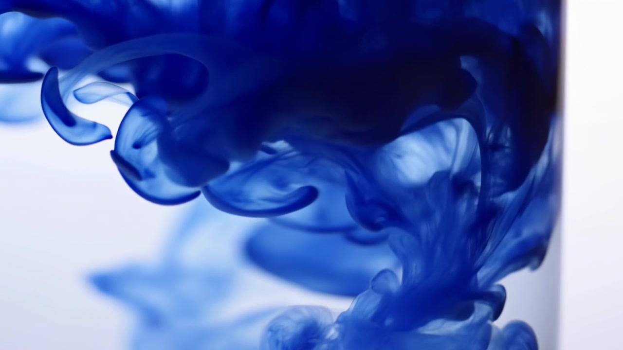 Detailní dynamická scéna koncentrovaného modrého inkoustu vstupujícího do čisté vody uvnitř průhledné nádoby, šířícího se do hustých oblaků, kroutících se stuh a vlečných vláken, zatímco bledne do jemných pramínků.