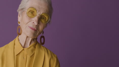 anciana de pelo corto con camisa y chaqueta de color mostaza, pendientes y gafas de sol posando y mirando la cámara con fondo morado