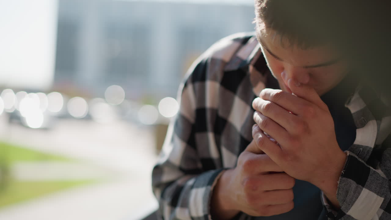 close up of smoker in black white plaid shirt holding cigarette between fingers cups hand around tip shields flame from breeze sparks ember against blurred cityscape background sunlit scene