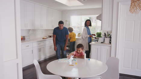 Papá-Viendo-A-Su-Hijo-Pequeño-Pintando-En-La-Cocina,-La-Madre-Y-La-Hija-Preparan-La-Comida-En-El-Fondo