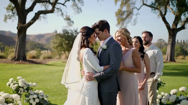 Vows beginning, leaning bride and groom embracing on sunny lawn, veil, gown and gray suit
