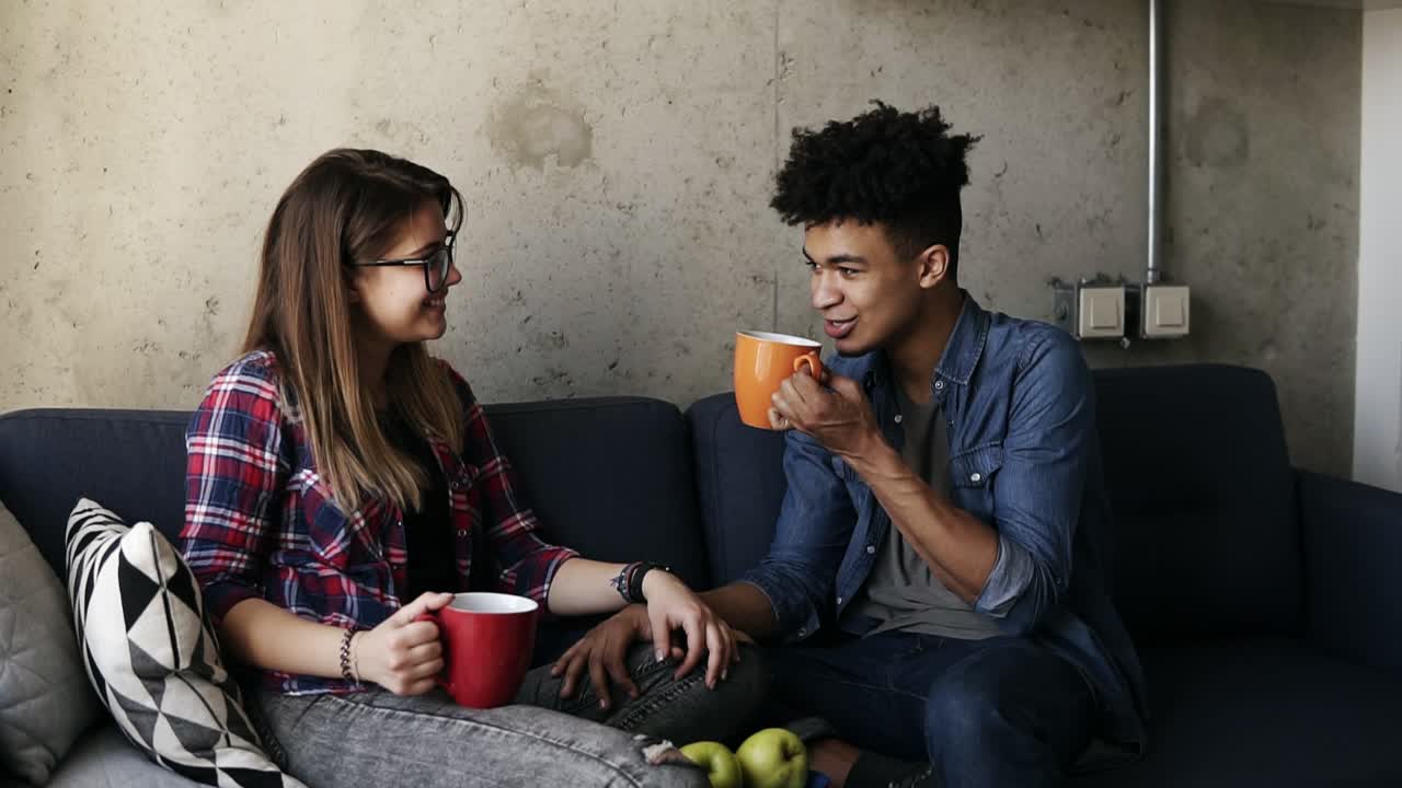una joven caucásica y un apuesto mulato, ambos con trajes urbanos, sentados en el sofá con tazas de té, hablando de la vida, disfrutando de pasar tiempo juntos.