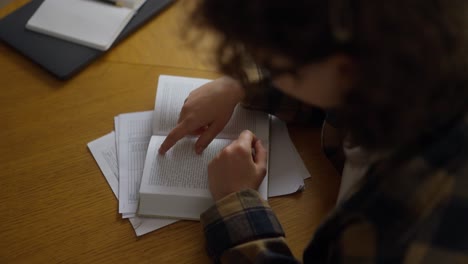 over the shoulder, a concentrated girl student with curly hair reads a book while sitting at a table in the library