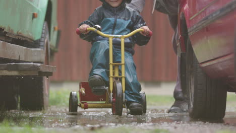 SLOW-MOTION,-FUNNY-shot-of-a-child-riding-a-colourful-trike-through-a-muddy-puddle