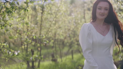 stunning brunette woman in white dress is enjoying walk in blooming garden whirling between trees