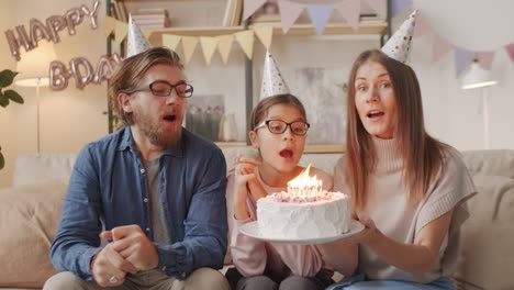 Padres-E-Hija-Celebrando-El-Cumpleaños-Con-Sombreros-De-Cumpleaños,-La-Niña-Sopla-Las-Velas-Del-Pastel-De-Cumpleaños