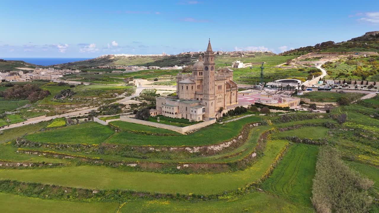 Un'inquadratura aerea con drone mostra il Santwarju Bazilika tal-Madonna ta' Pinu sullo sfondo di lussureggianti campi terrazzati verdi e della costa mediterranea a Gozo, Malta.