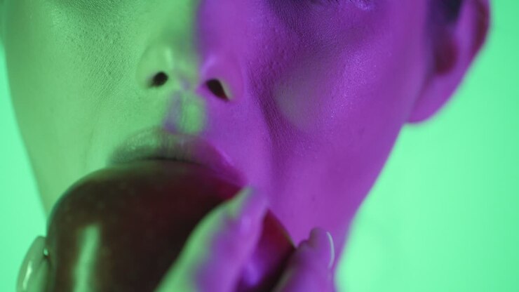 Extreme close up of a woman lip while she takes a pleasurable bite of a red apple in front of green background with green and purple contrast in her beautiful face in slow motion