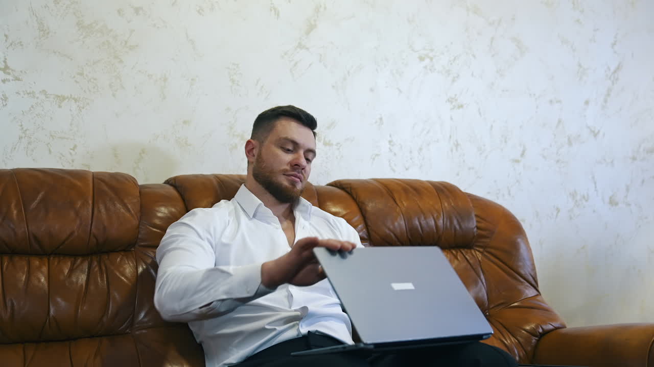 Freelancer using laptop while sitting on sofa. Tired businessman works online in business trip. Man resting on couch in hotel room. Business relaxing concept.