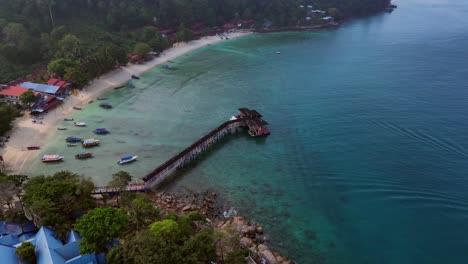 Morning-mood-Aerial-view-speed-ramp-of-a-tropical-island-with-a-long-wooden-pier-jetty-leading-to-a-floating-restaurant,-surrounded-by-turquoise-waters-and-lush-green-rainforest