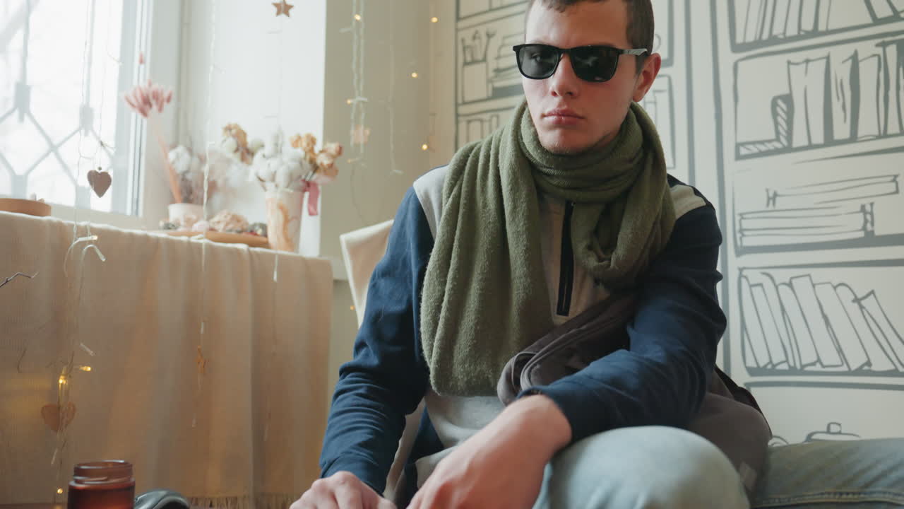 White man wearing sunglasses at laptop in airy interior, green scarf and denim, poised sitting posture with steady hands, layered backdrop of illustrated bookshelf and dried flowers, sequence shifts