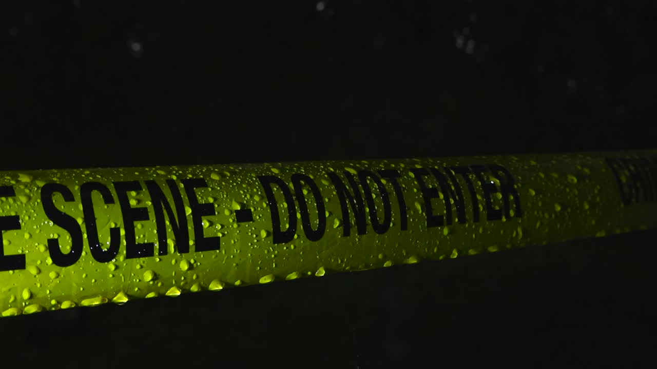 Close up footage of a yellow police line forensics investigation Crime Scene tape covered in raindrops and waterdrops during a rainy dark night, lit by good spotlight light from the side, slow motion