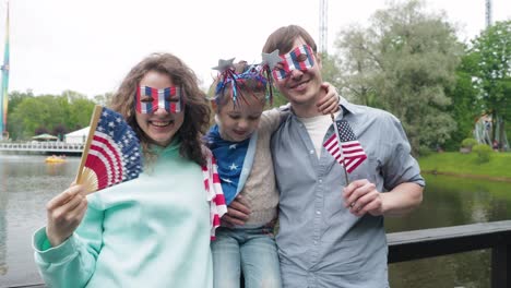 portrait of happy young family celebrating 4th july independence day holding silhouette usa national flag against park, parents kissing daughter. front view