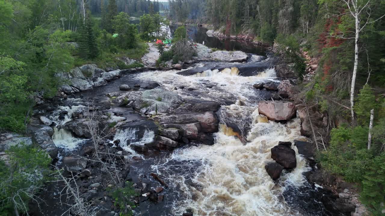 Manitoba'daki Manigotagan Nehri üzerinde, Kanada Kalkanı graniti üzerinden akan Turtle Şelaleleri. Havadan çekim yapan kamera, yaz aylarında nehir kenarında kamp ateşi ve hafif dumanla çadır ve kampçılara doğru yukarı yönde hareket ediyor.