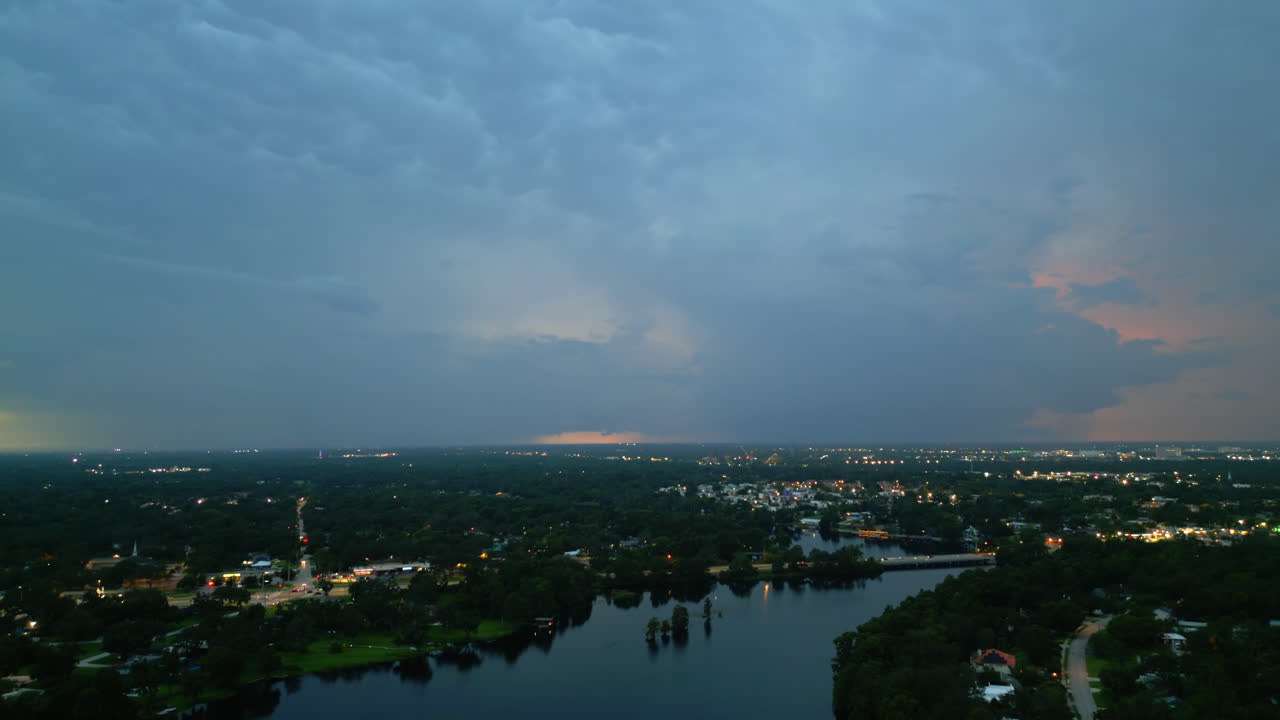 Aerial flying over a river reflecting the evening twilight sky as lightning flashes in the distance and the city skyline as soft ambient lights are turning on