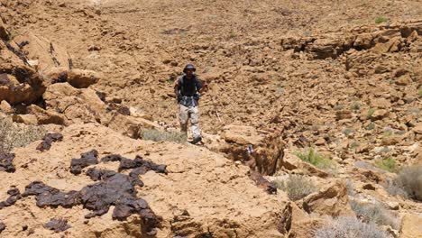 One-man-trekker-with-trekking-poles-walking-down-the-sandstone-cliffy-slope-during-travel-in-Ramon-Crater-Trail,-Negev-desert,-Israel