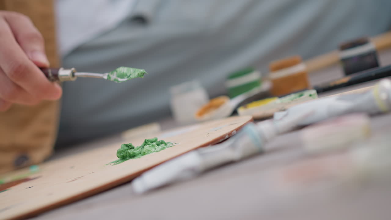 Nærbillede af hånd, der påfører impasto grøn maling med paletkniv, studieinstruktør demonstrerer tyk teksturteknik til workshop, palet og tuber arrangeret på træplade, pensler sløret