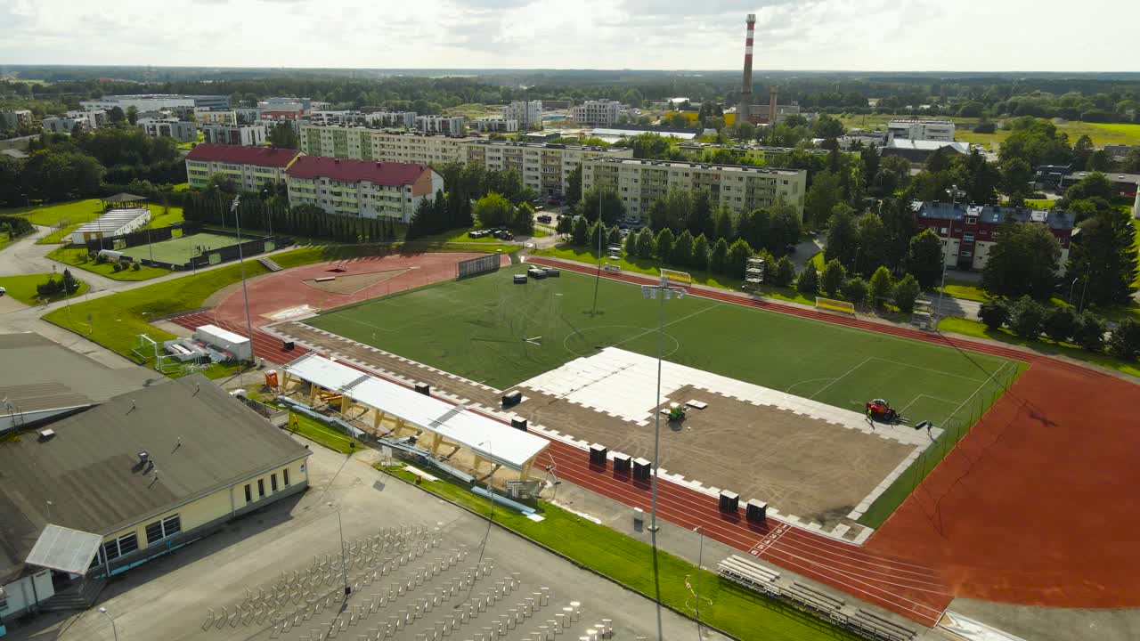 Aerial drone video flying over and orbiting around a large sunny summer green soccer stadium or field while it is being constructed or renovated by workers with machines, apartment buildings visible