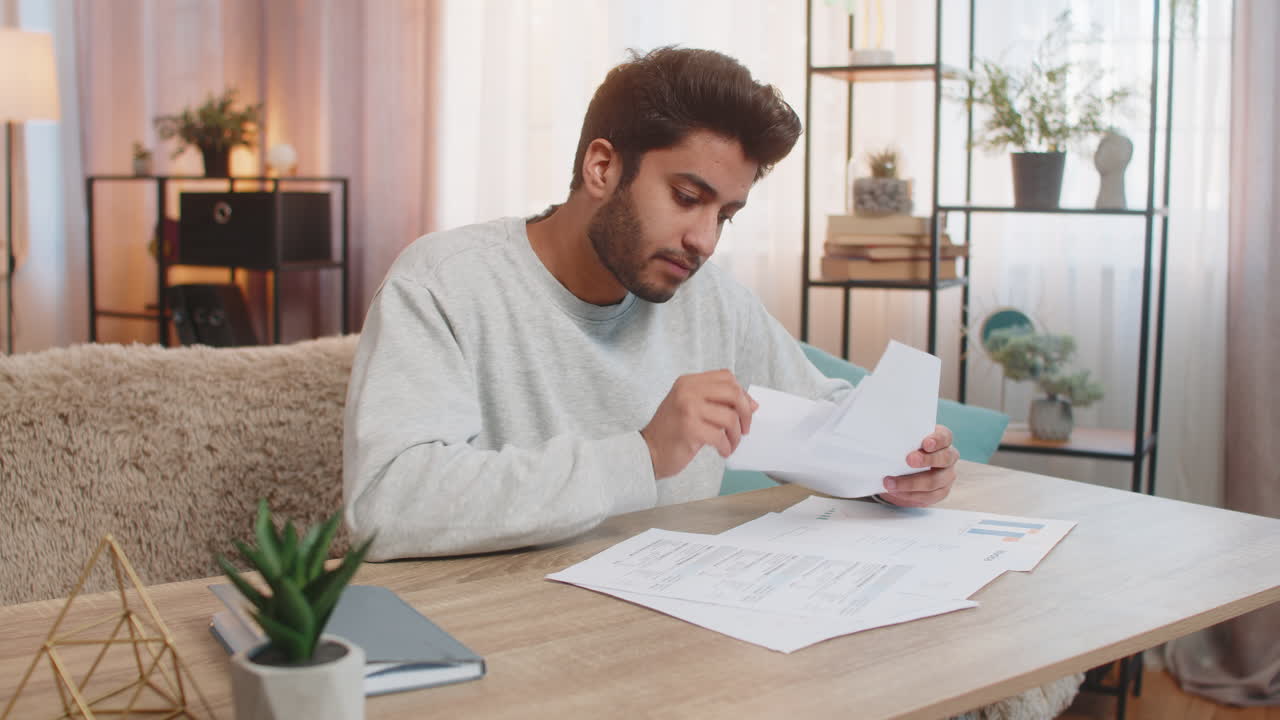 Young indian man frustrated with bills and expenses while reviewing documents at home on table