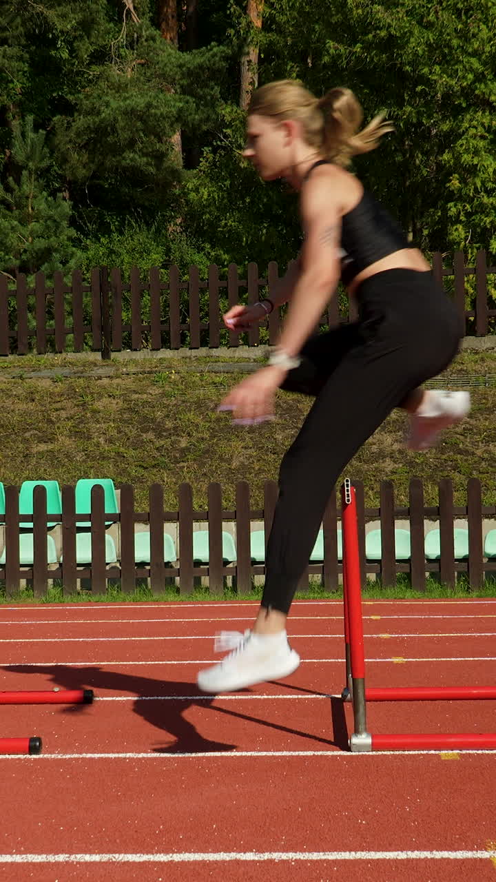 A fit young woman in black sportswear performs hurdle jumps on a red tartan track during a sunny outdoor training session. Perfect for fitness and sports content