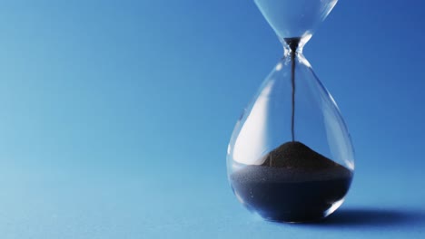 video of close up of hourglass with black sand pouring with copy space on blue background