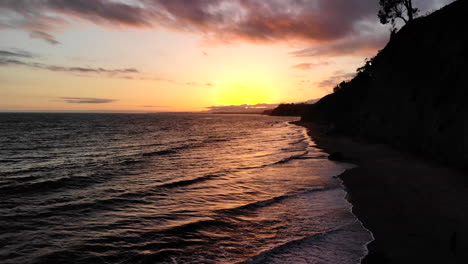 Aerial-drone-shot-of-a-sunset-sky-with-reflections-on-the-ocean-waves-near-the-coastline-cliffs-of-Santa-Barbara,-California