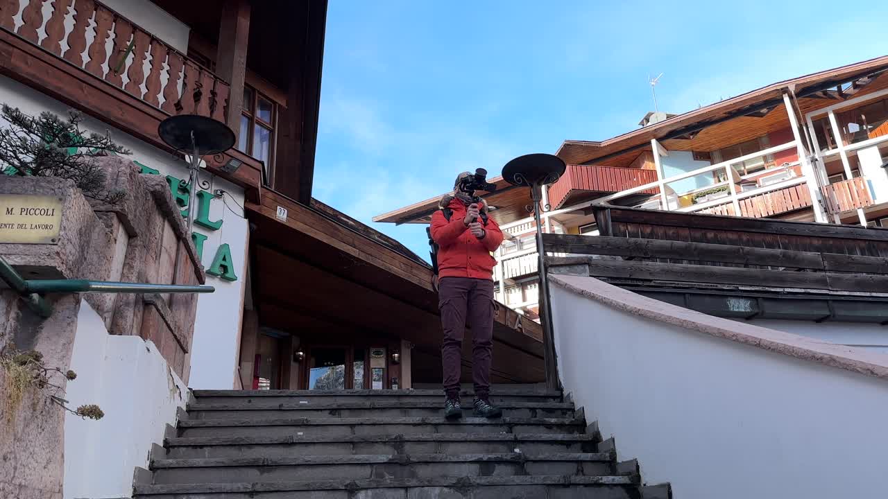 Travel Photographer With Camera On Gimbal Stabilizer Standing On Stairs, Taking Photos In Cortina d'Ampezzo, Italy
