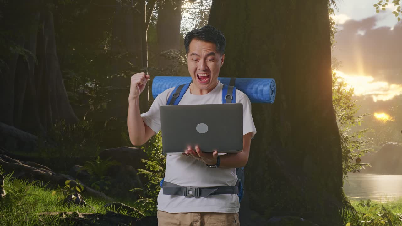 산악 배을 가진 아시아 남성 하이커가 노트북을 보고 숲의 자연을 탐험하는 동안 성공을 축하하는 목표를 외치고 있습니다.