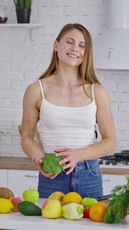 cheerful female with broccoli at home. sportive young woman choosing healthy diet full of minerals and vitamins in the kitchen. girl standing near fresh fruit and vegetables. vertical video